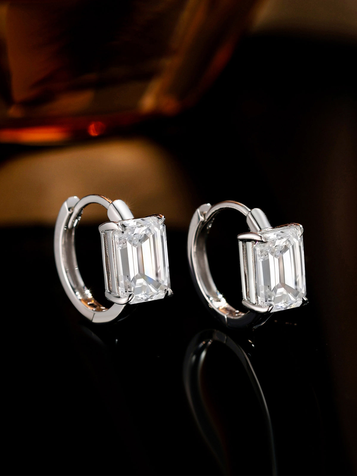Emerald-cut diamond huggie earrings on a dark reflective surface