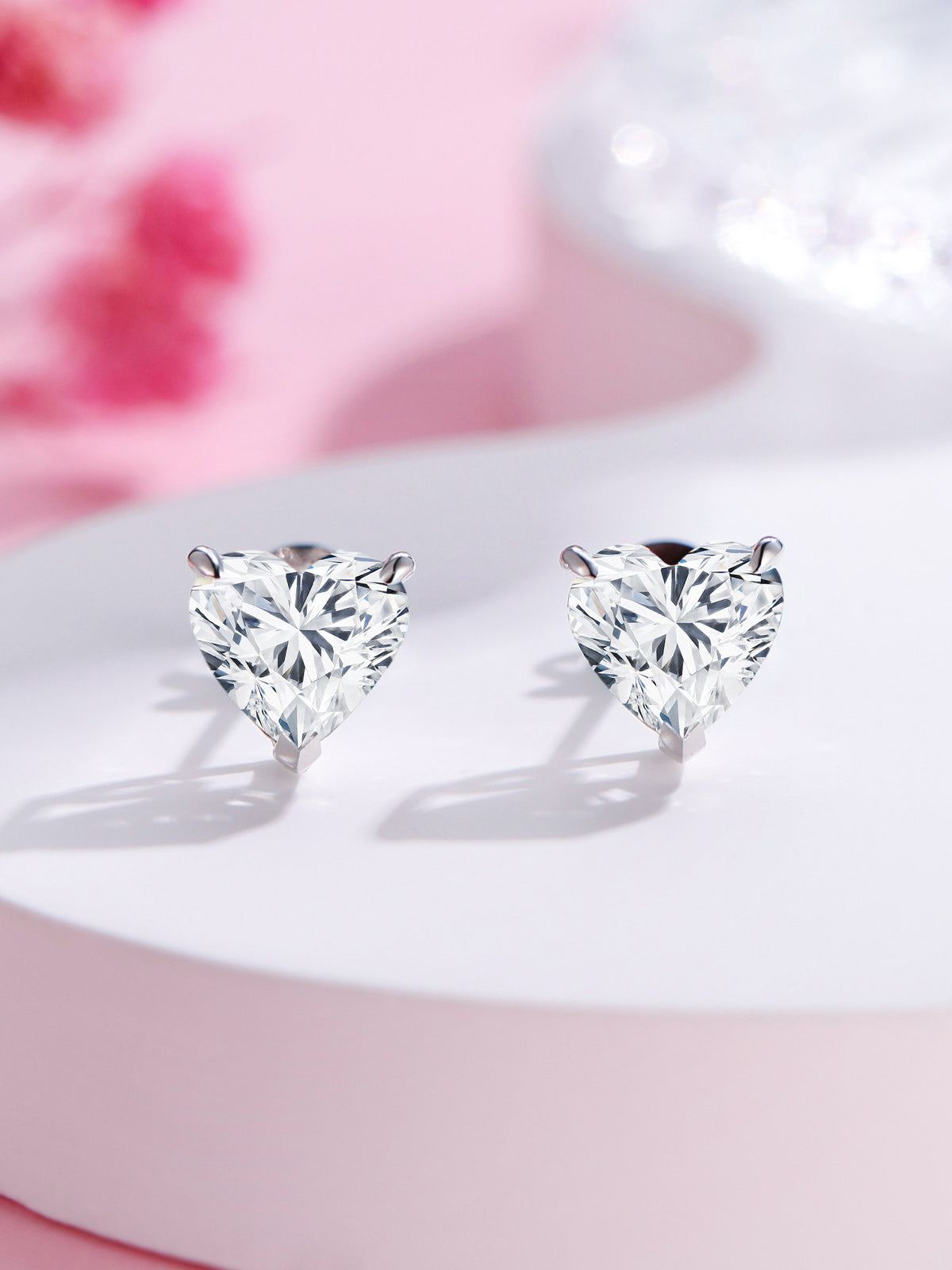 Three-prong heart-cut lab-grown diamond stud earrings on a pink backdrop