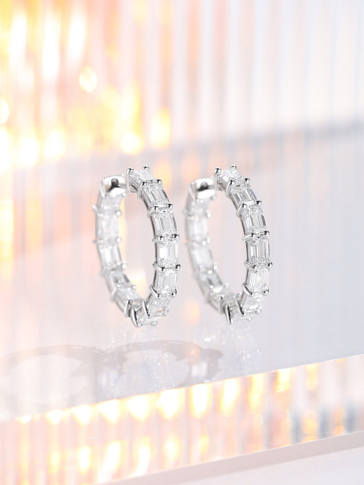 Pair of emerald-cut lab-grown diamond hoop earrings in platinum standing on acrylic with warm light