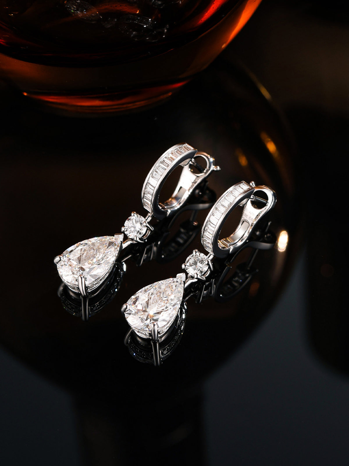 Pear-cut lab-grown diamond drop earrings with baguette huggies on a dark mirror beside amber glass